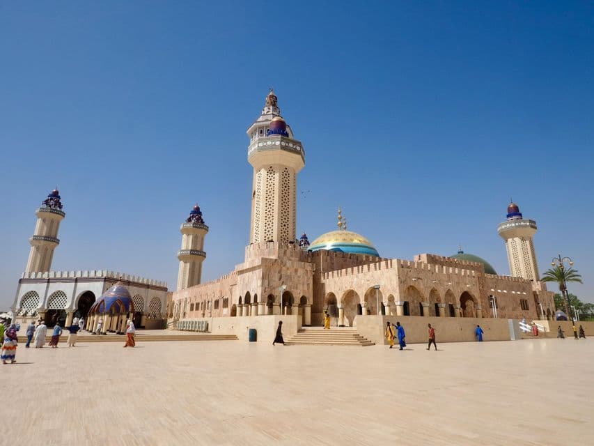 Una mezquita de piedra ornamentada con altos minaretes y cúpulas coloridas se alza bajo un cielo azul claro mientras la gente camina por su gran plaza.