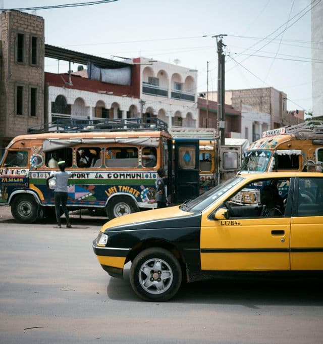 Un taxi amarillo y negro en una carretera pavimentada con un autobús colorido y otros vehículos al fondo.