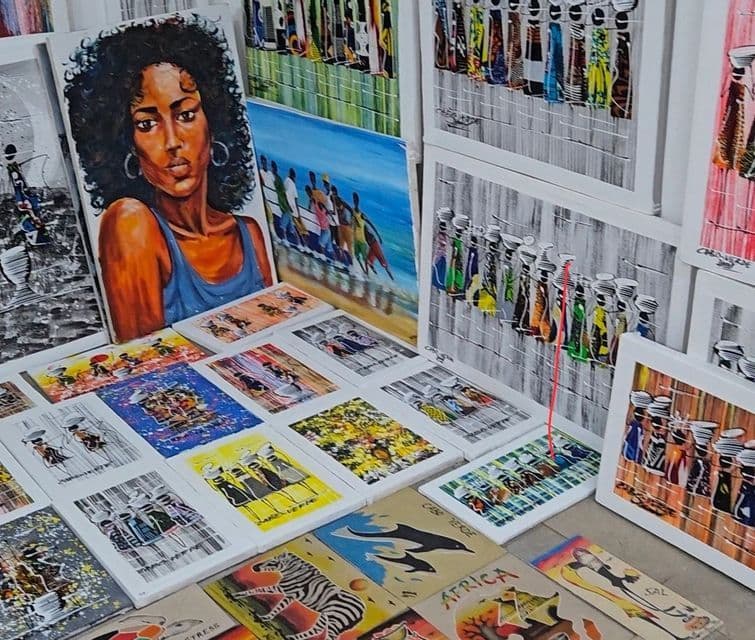 Eine Sammlung farbenfroher Leinwandbilder, darunter ein Frauenporträt, die afrikanische Motive zeigen und an einem Marktstand präsentiert werden.