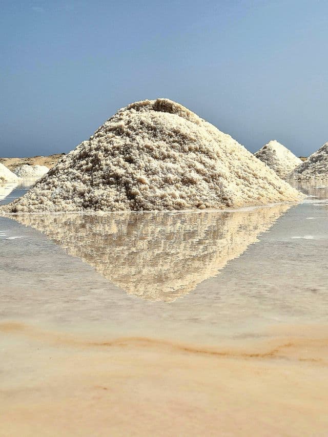 Ein großer Haufen groben weißen Salzes spiegelt sich im stillen, flachen Wasser einer Salzpfanne unter klarem Himmel.