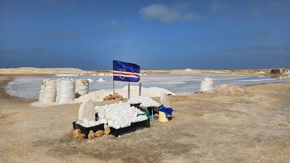 The flag of Cape Verde stands amongst piles of harvested salt in bags and crates at a salt pan under a blue sky.