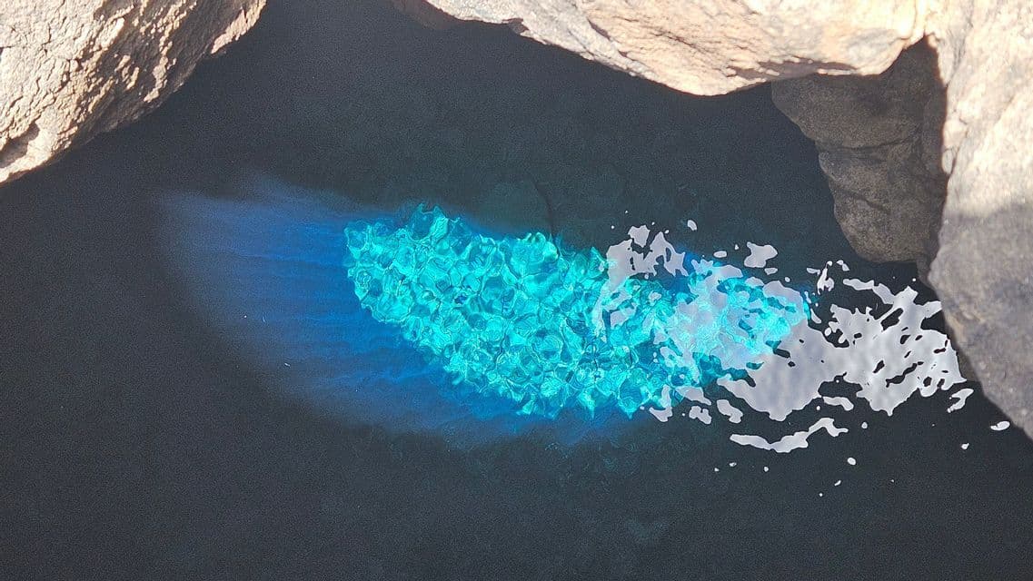 Sunlight creates a bright turquoise patch on the surface of dark water inside a rocky cave.