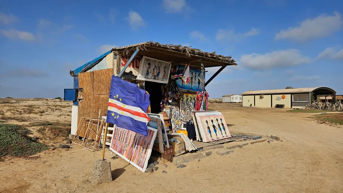 A rustic souvenir stall displays paintings and a flag with a circle of stars in a sandy, arid landscape.