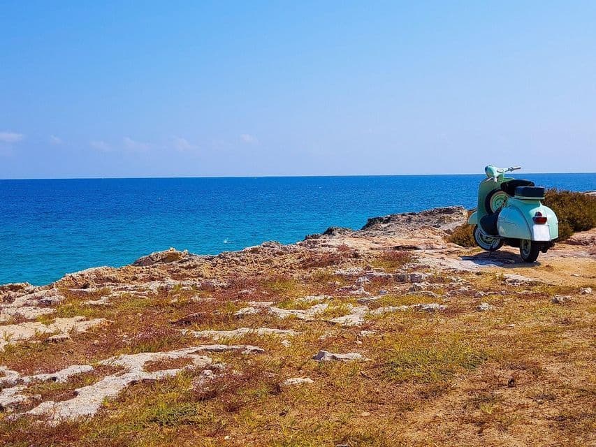 Uno scooter vintage azzurro parcheggiato su una scogliera rocciosa, con vista su un mare calmo e azzurro e un cielo sereno.