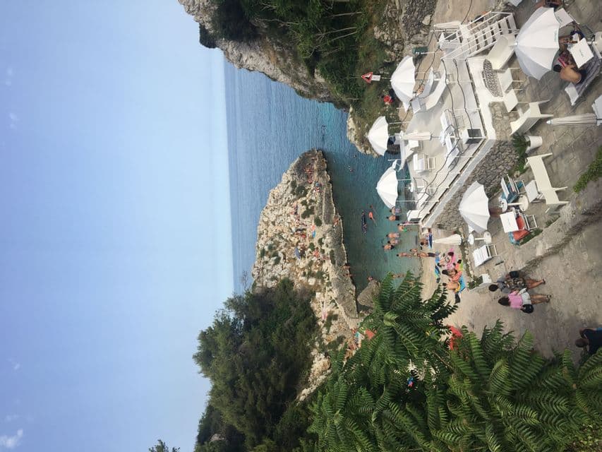 Vista dall'alto di una caletta rocciosa dove le persone nuotano e prendono il sole vicino a una terrazza su scogliera bianca con ombrelloni.