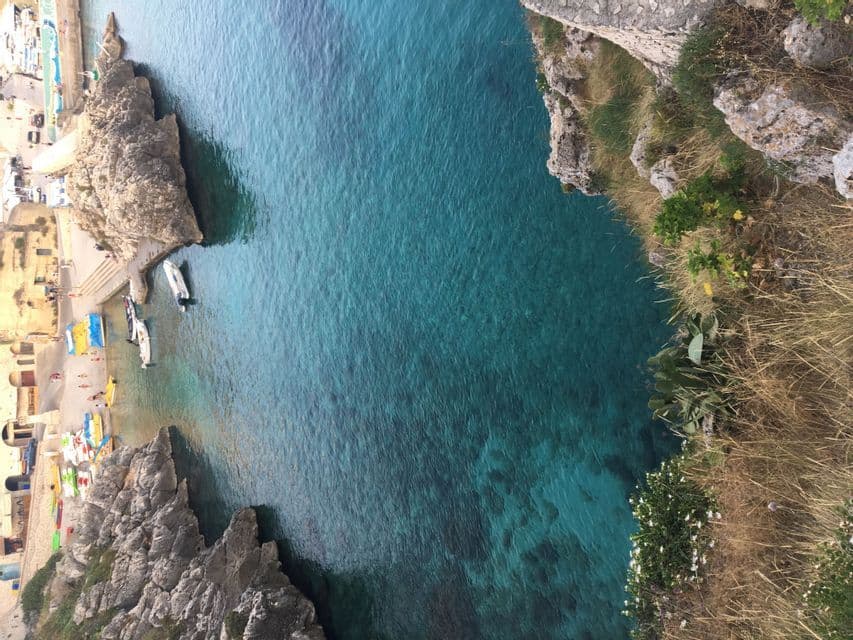 Vista dall'alto di una cala rocciosa con acqua limpida e turchese, una spiaggetta e diverse barche ormeggiate.