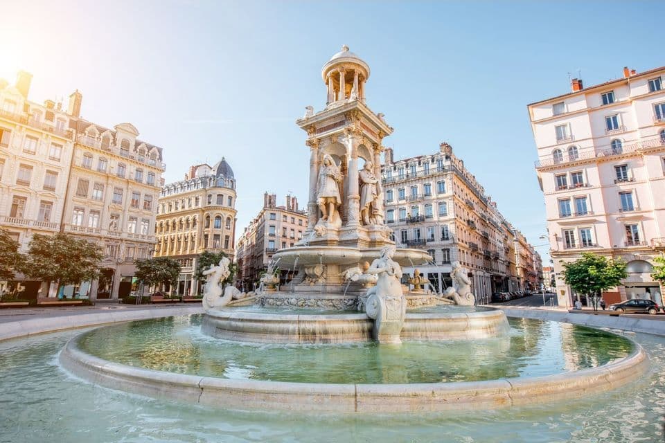Une fontaine en pierre ornée de sculptures au centre d'une place de ville, entourée de bâtiments européens historiques par une journée ensoleillée.