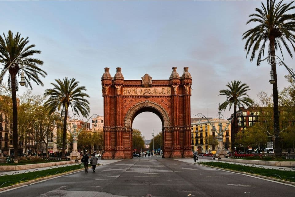 An ornate red brick triumphal arch stands at the center of a wide promenade flanked by tall palm trees at dusk.