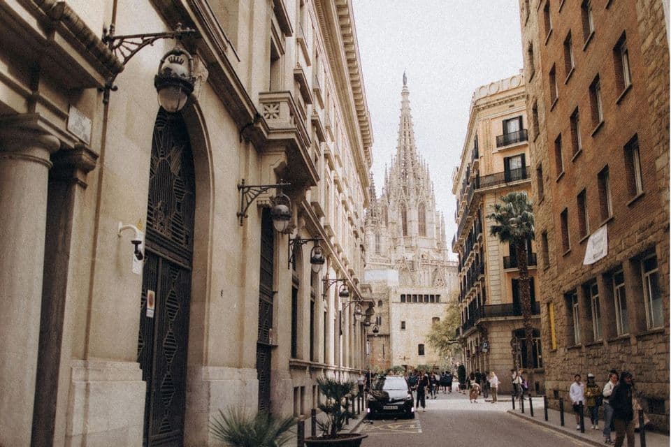 A narrow city street flanked by historic buildings, with the ornate spires of a Gothic cathedral visible in the distance.