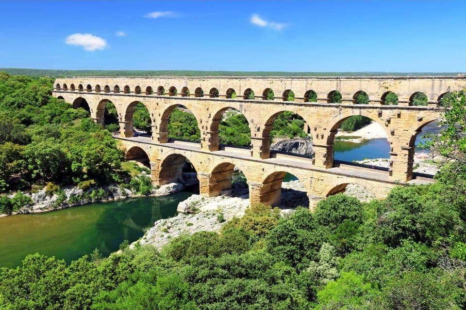 A three-tiered stone aqueduct with arches spans a river, surrounded by lush green forest under a bright blue sky.