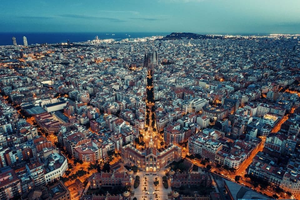 Una vista aérea de un vasto paisaje urbano iluminado al anochecer, extendiéndose hacia el mar bajo un cielo azul profundo.