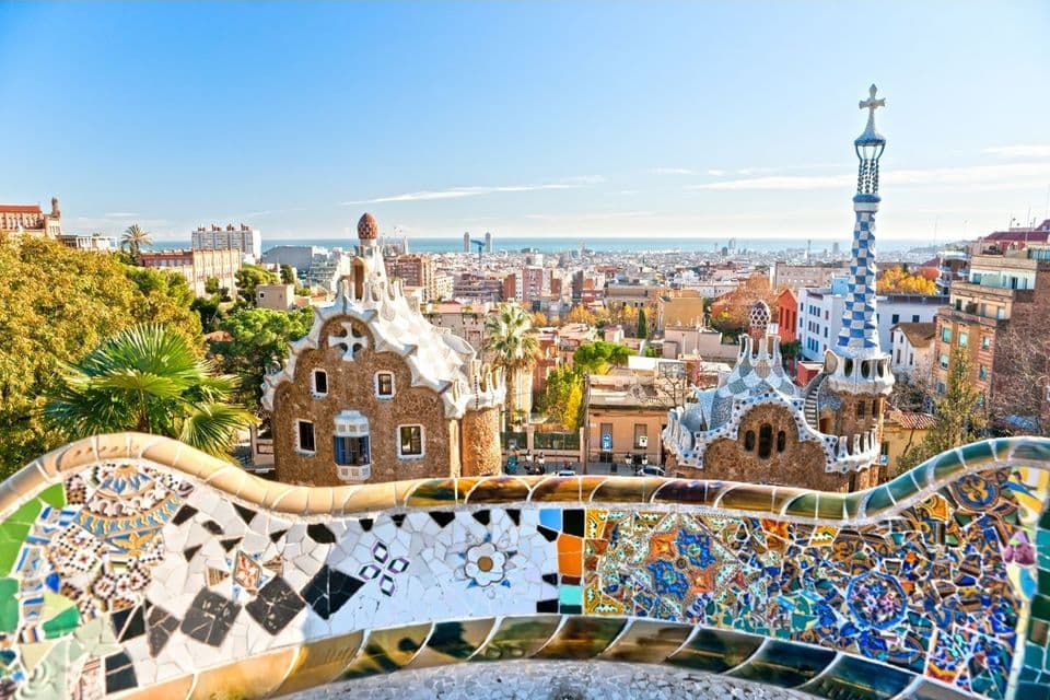 Un banco de mosaico colorido con vistas a edificios caprichosos y un paisaje urbano que se extiende hasta el mar bajo un cielo azul claro.