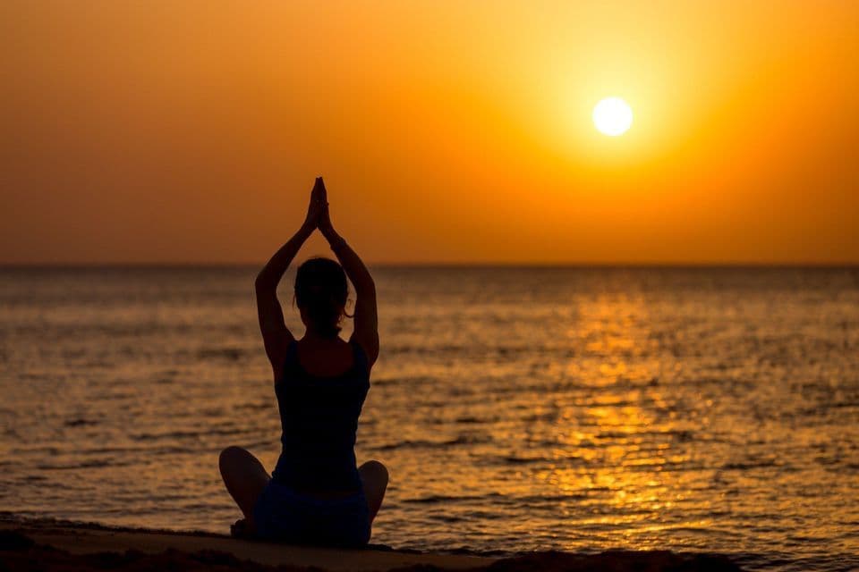 Une personne en pose de yoga sur une plage, en silhouette devant un coucher de soleil orange sur l'océan.