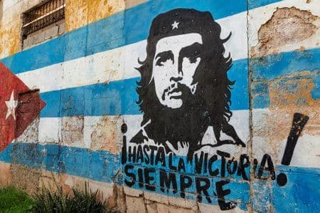 Ein Street-Art-Wandbild auf einer verwitterten Mauer, das Che Guevara über einer kubanischen Flagge mit dem Spruch 'Hasta la Victoria Siempre' zeigt.