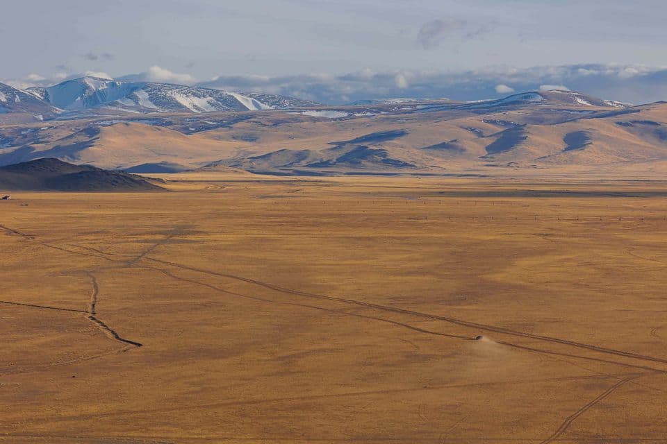 Un véhicule solitaire traverse une vaste steppe dorée, ses traces de pneus menant vers des montagnes enneigées au loin, sous un ciel nuageux.