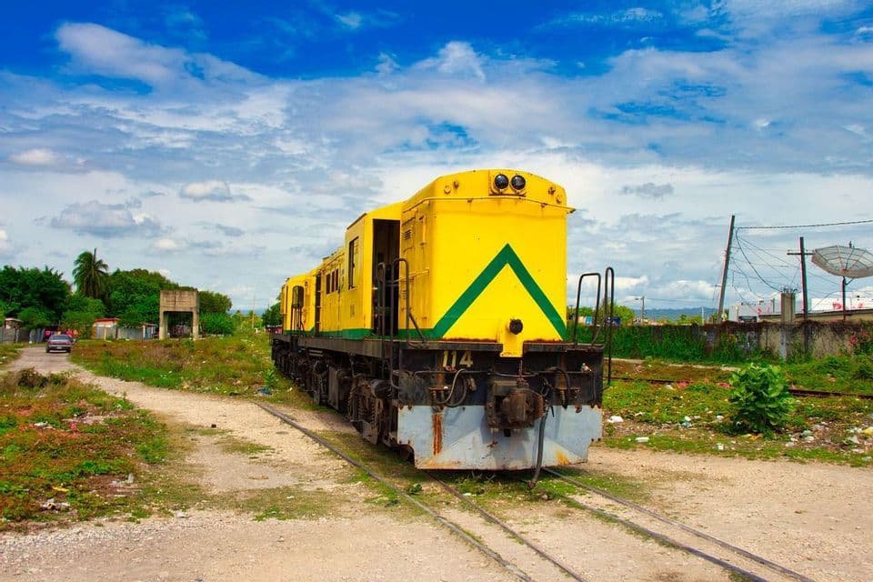 Un train jaune vif à bande verte est à l'arrêt sur une voie ferrée dans une zone rurale, sous un ciel bleu avec des nuages blancs.