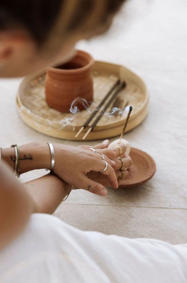 Primo piano delle mani di una persona con anelli e tatuaggi che tengono un bastoncino d'incenso fumante in un piatto di terracotta.