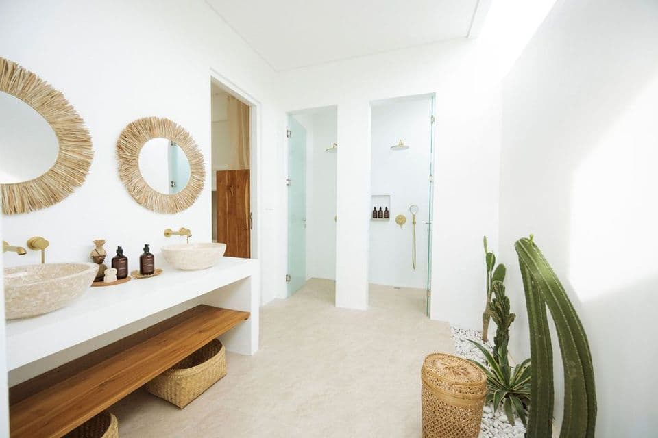 Un bagno bianco minimalista con due lavandini in pietra, specchi con cornici in paglia e docce separate in vetro. Una pianta di cactus si trova nell'angolo.