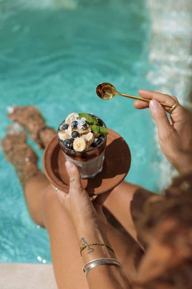 Una persona tiene in mano un bicchiere di budino di chia guarnito con frutta, seduta con i piedi in piscina.