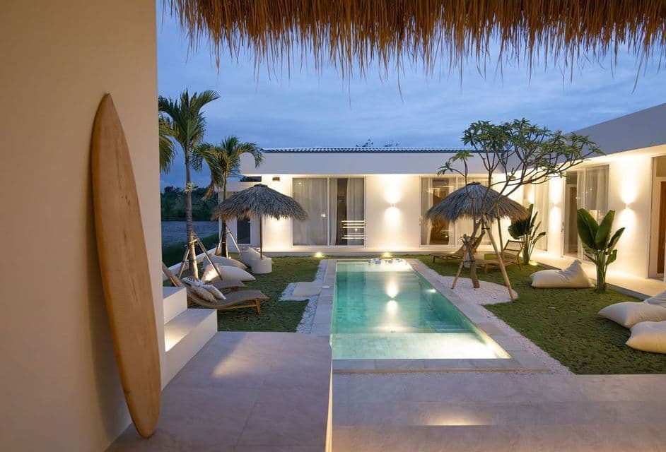Un moderno cortile di villa al tramonto, con piscina illuminata, ombrelloni in paglia e una tavola da surf appoggiata al muro.