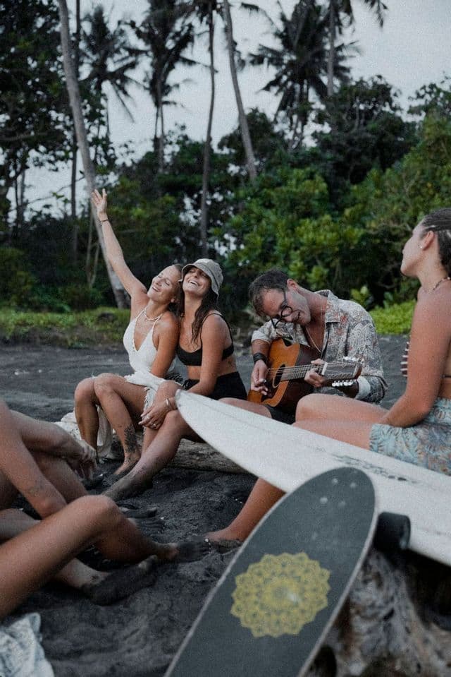 Un gruppo WeRoad seduto su un tronco su una spiaggia di sabbia scura, con una persona che suona la chitarra acustica.