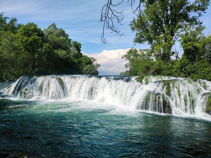 Une grande cascade s'écoule sur des rochers moussus dans une rivière turquoise, entourée d'arbres luxuriants.