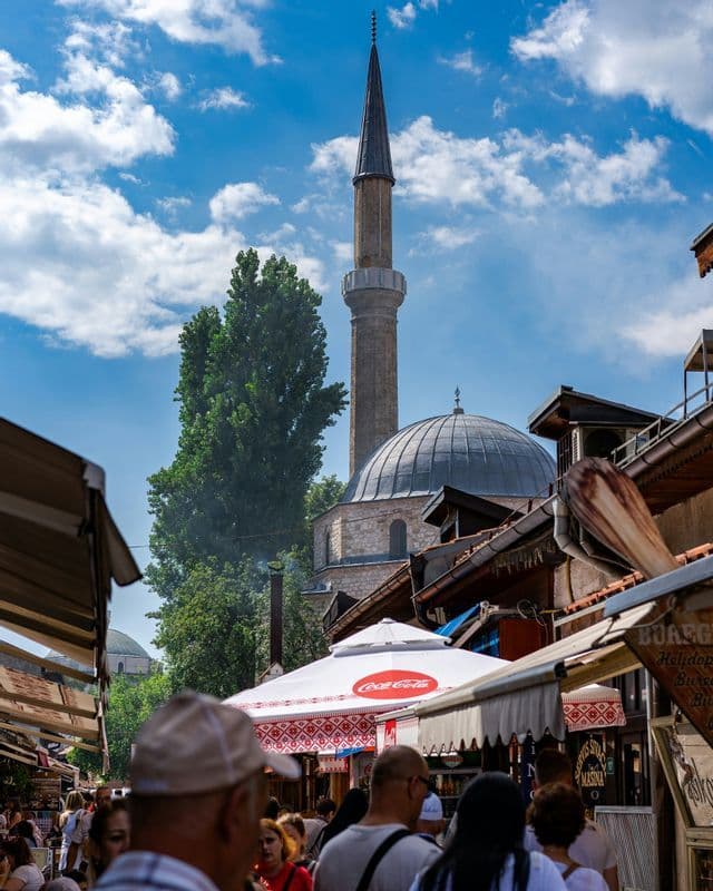 Une foule traverse une rue de marché animée, avec une mosquée en pierre et un minaret se dressant sous un ciel bleu nuageux.