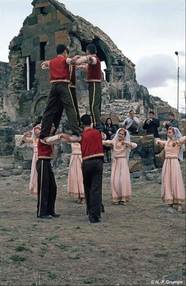 Vor steinernen Ruinen führt eine Gruppe in traditioneller Tracht einen Volkstanz auf und bildet eine menschliche Pyramide.