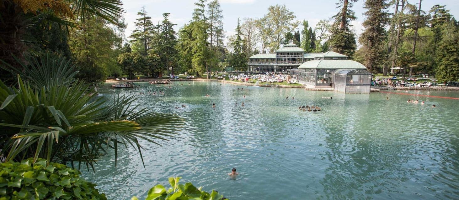 Persone che nuotano e si rilassano in un ampio lago termale, circondato da alberi verdi lussureggianti e un edificio in vetro sulla riva.