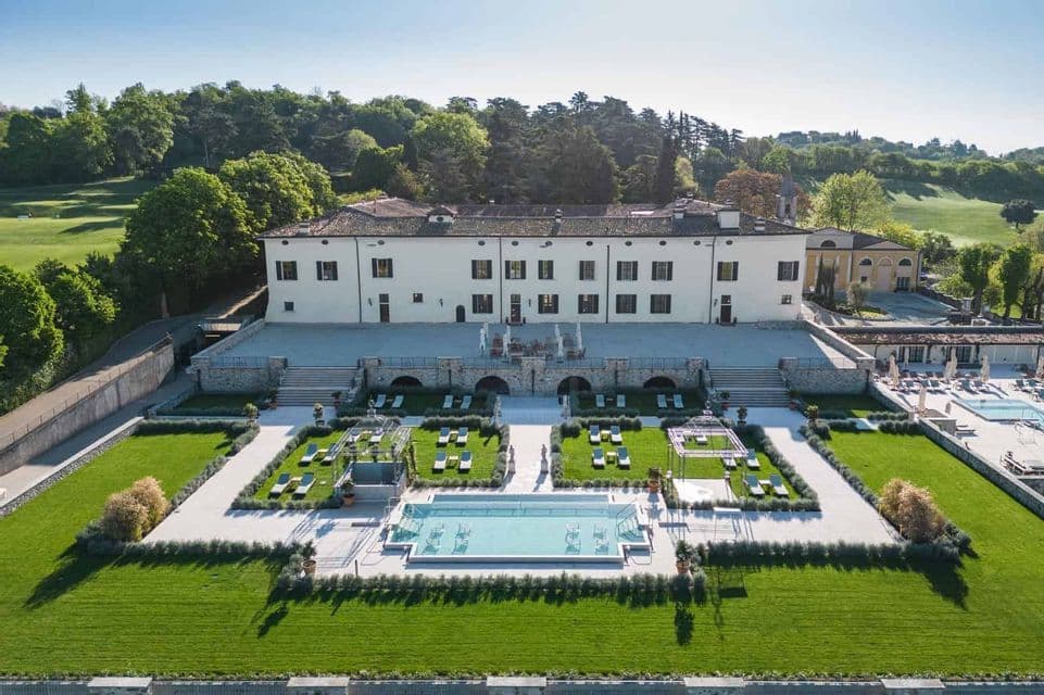 Veduta aerea di un'ampia villa bianca con un giardino formale, piscina e lettini su un prato curato.