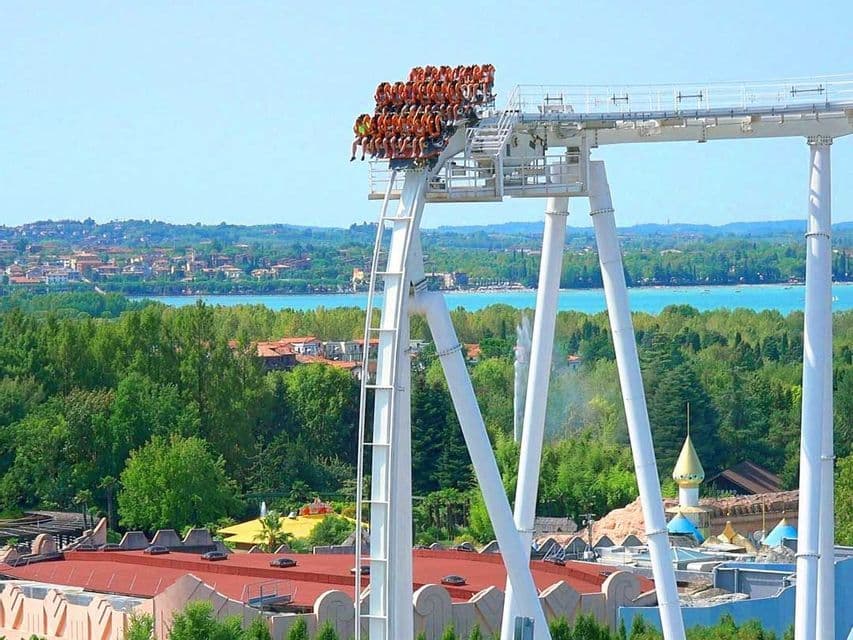 Un vagone delle montagne russe, pieno di persone, si è fermato in cima a una ripida discesa, affacciandosi su un grande lago e un paesaggio verde.