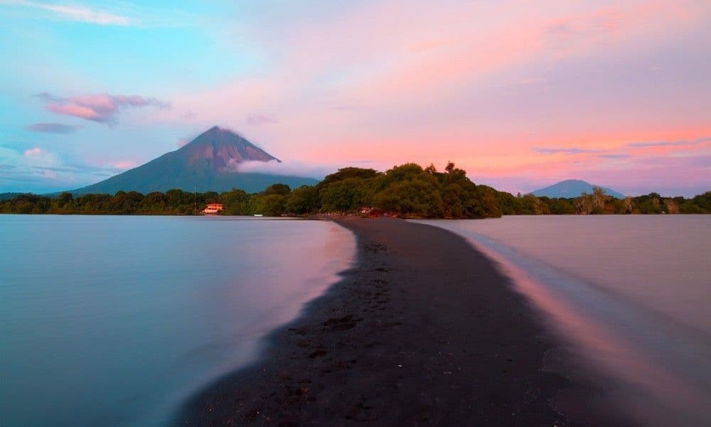 Una lingua di sabbia nera si curva in un lago calmo con un grande vulcano sullo sfondo sotto un cielo al tramonto rosa e blu.