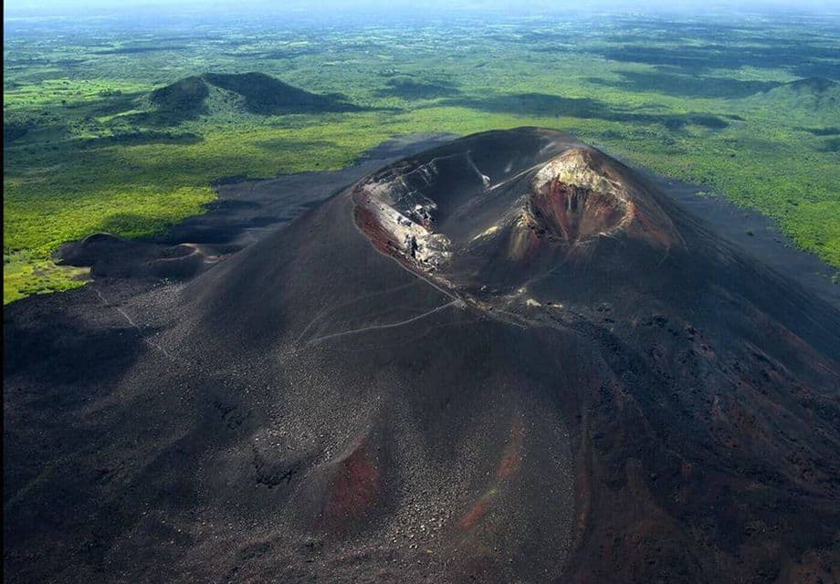 Vista aerea di escursionisti su un sentiero che sale lungo il fianco di un grande vulcano scuro, in netto contrasto con il lussureggiante paesaggio verde sottostante.