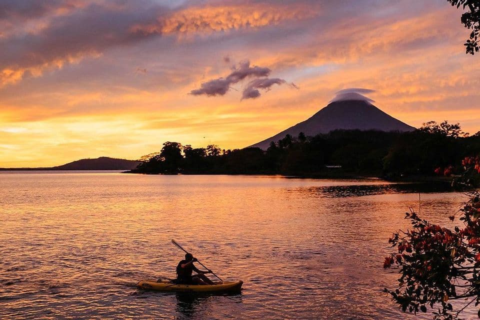 Una persona in kayak su un lago al tramonto, con un vulcano in silhouette sotto un cielo colorato.