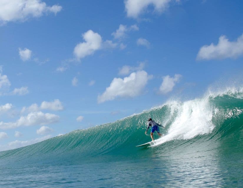 Un surfista in pantaloncini blu e maglietta a maniche lunghe cavalca un'onda turchese grande e ricurva sotto un cielo parzialmente nuvoloso.