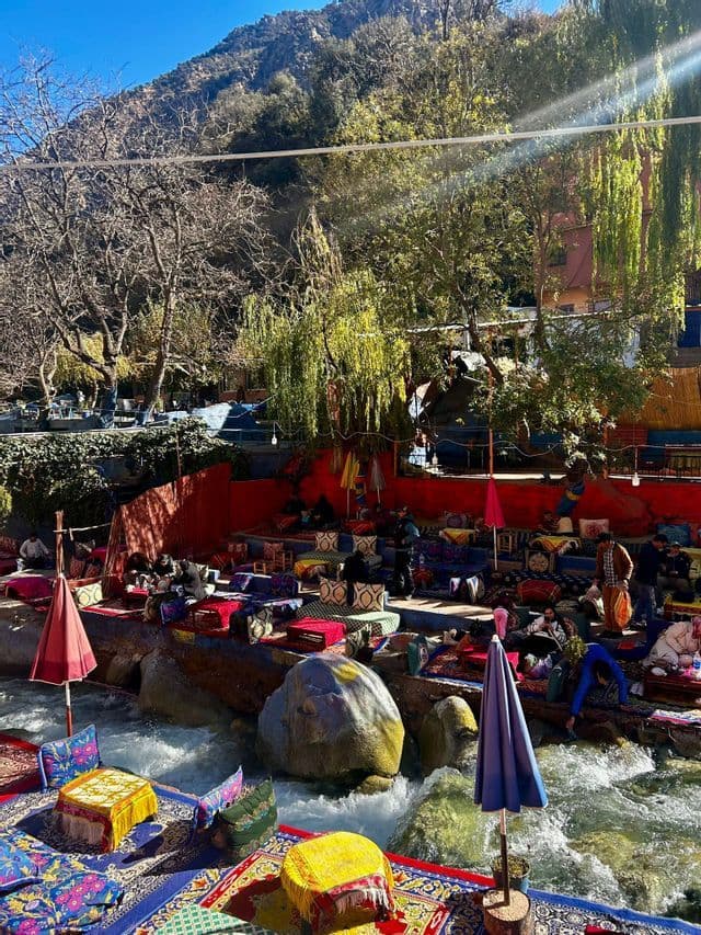 Un colorido café al aire libre con asientos sobre alfombras y cojines dispuestos en rocas, junto a un río caudaloso en un paraje montañoso y boscoso.