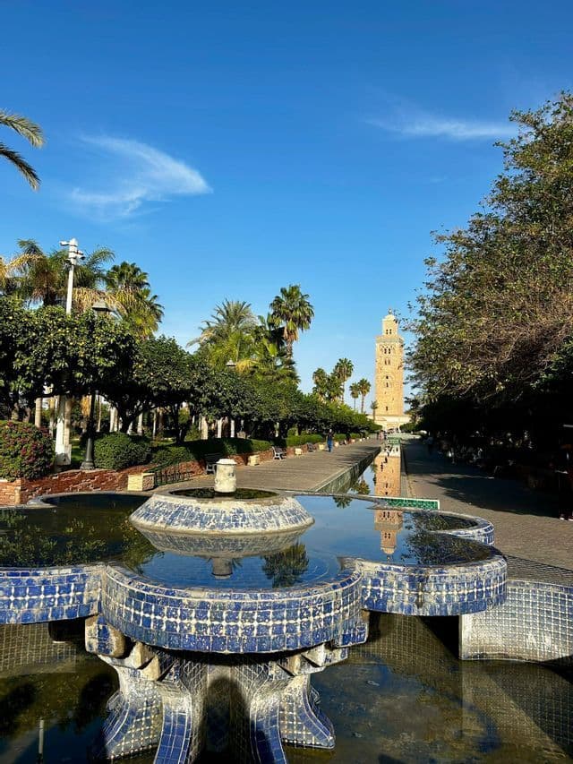 Una fuente de mosaico azul en un parque refleja una torre alta al final de un largo paseo bordeado de árboles.