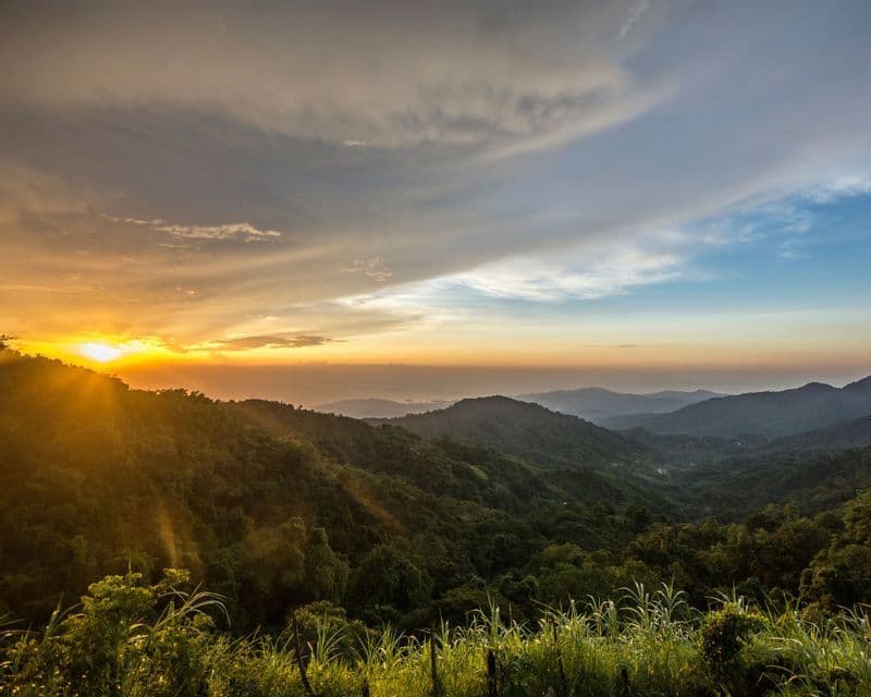 Die Sonne geht über einem Tal üppig grüner Berge unter und wirft goldene Sonnenstrahlen durch die Bäume unter einem teilweise bewölkten Himmel.