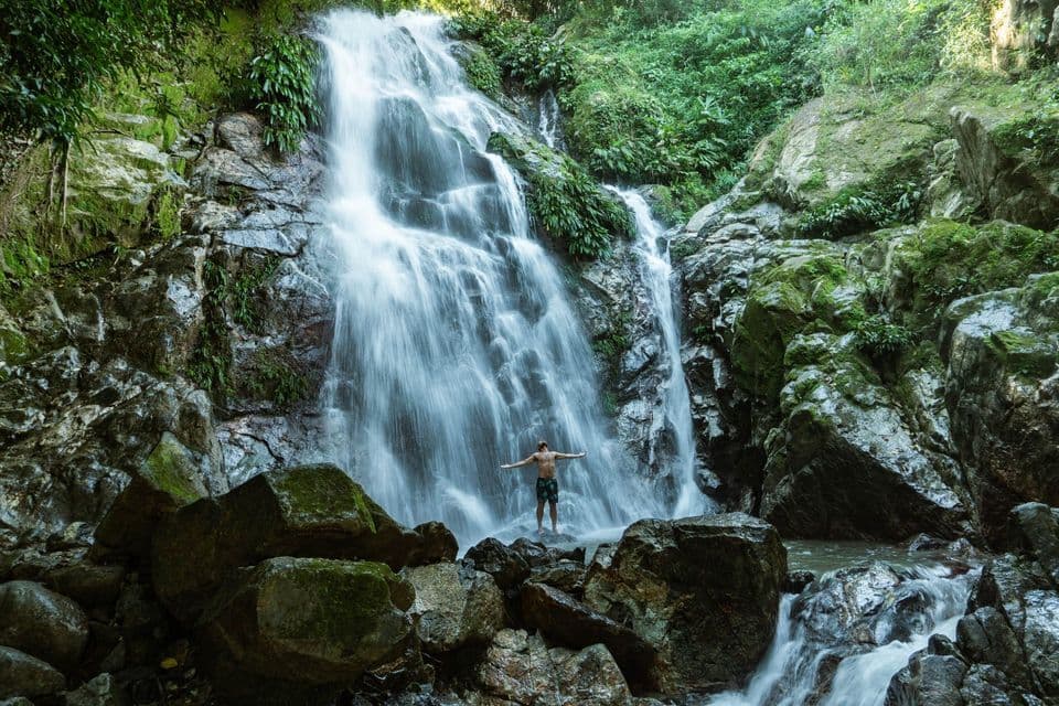 A shirtless man stands with his arms outstretched on rocks at the base of a cascading waterfall in a lush jungle.