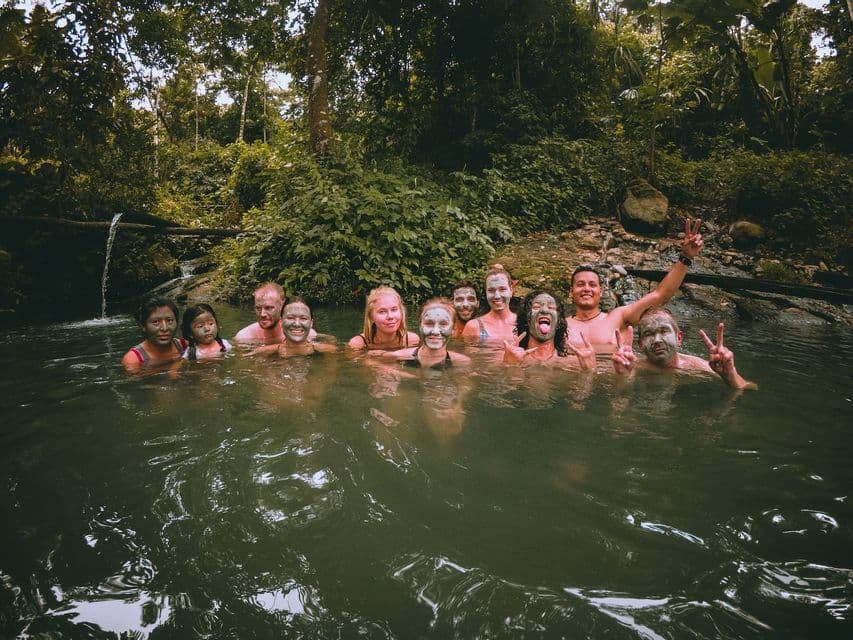 Un gruppo WeRoad sorride e posa in un fiume con il fango sui volti, circondato da una giungla lussureggiante.
