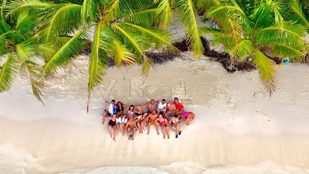 Vista aérea de un viaje en grupo de WeRoad posando en una playa de arena blanca bajo palmeras, con 'WeRoad' escrito en la arena.