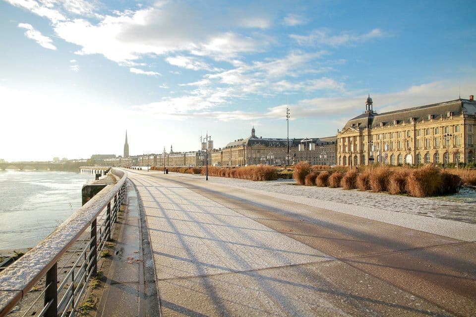 Un amplio paseo, cubierto de nieve, se extiende junto a un río, con una larga fila de edificios clásicos bajo un cielo azul y soleado.