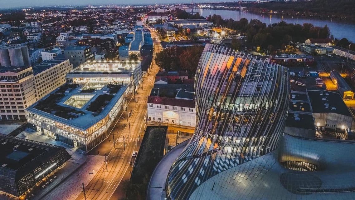 Vista aérea de un moderno edificio de cristal curvado, con vistas a una ciudad con calles iluminadas y un río al anochecer.