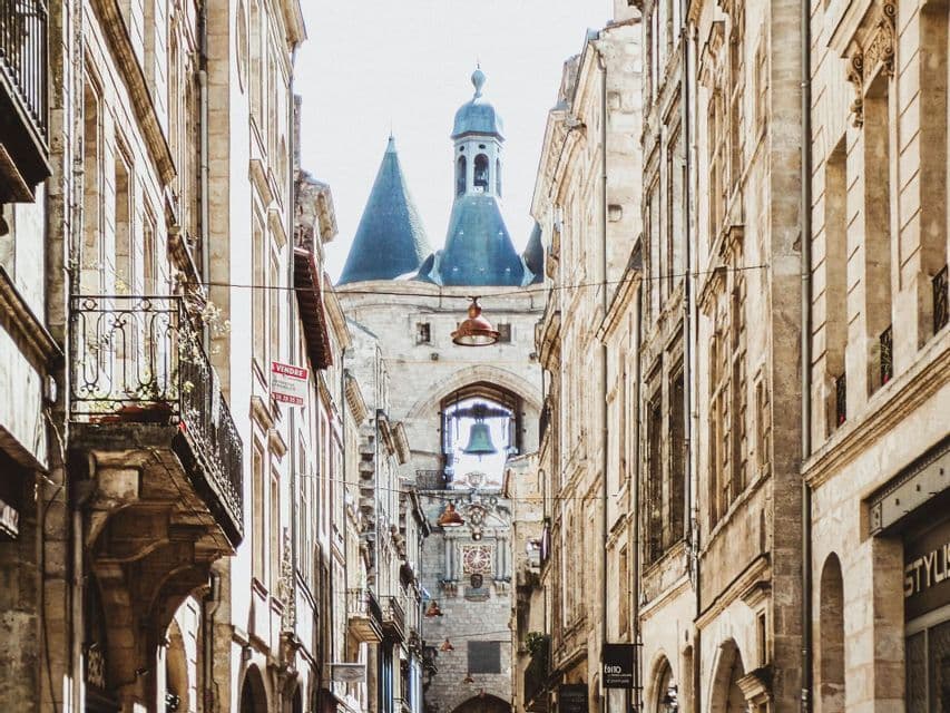Una calle europea estrecha flanqueada por edificios de piedra lleva a una gran torre del reloj con una campana visible en su arco.