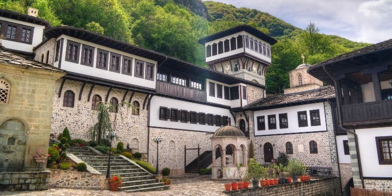 Historische Stein- und weiß getünchte Gebäude mit Holzbalkonen umgeben einen gepflasterten Innenhof am Fuße eines grünen Berges.