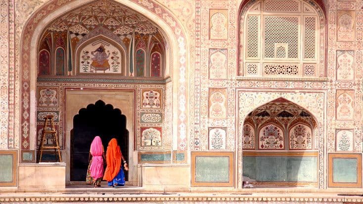 Tre donne in sari colorati attraversano un portale ad arco di una facciata di palazzo riccamente decorata in India.