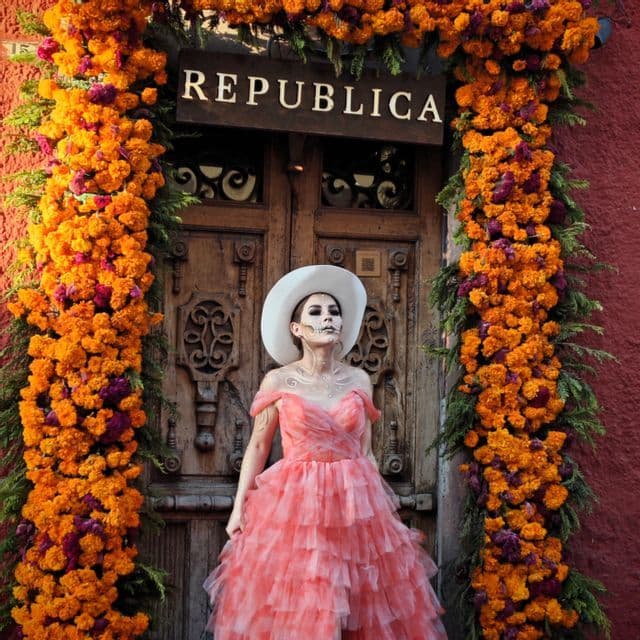Eine Frau mit Catrina-Make-up und einem rosa Kleid steht an einer mit orangefarbenen Ringelblumen geschmückten Holztür.