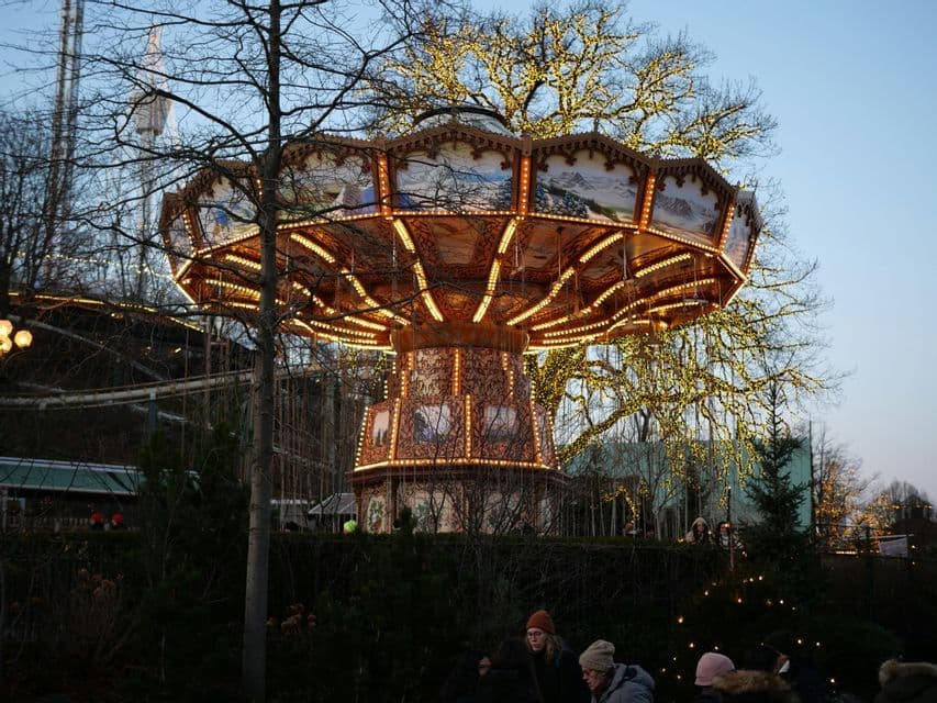 Ein beleuchtetes Kettenkarussell in einem Freizeitpark in der Abenddämmerung, umgeben von kahlen Bäumen, die mit Lichterketten geschmückt sind.