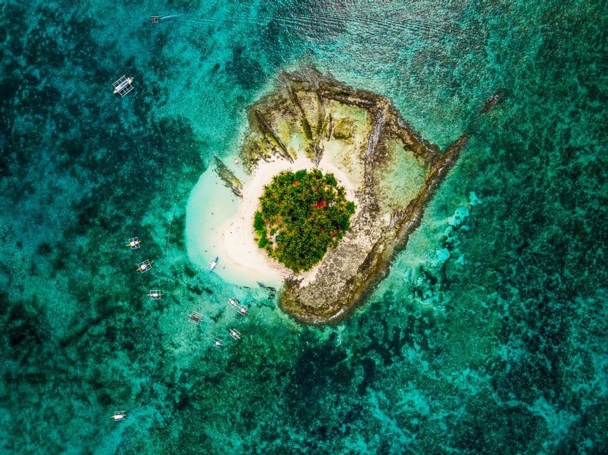 Vista aerea dall'alto di una piccola isola tropicale con palme, circondata da acqua turchese trasparente e diverse piccole imbarcazioni.