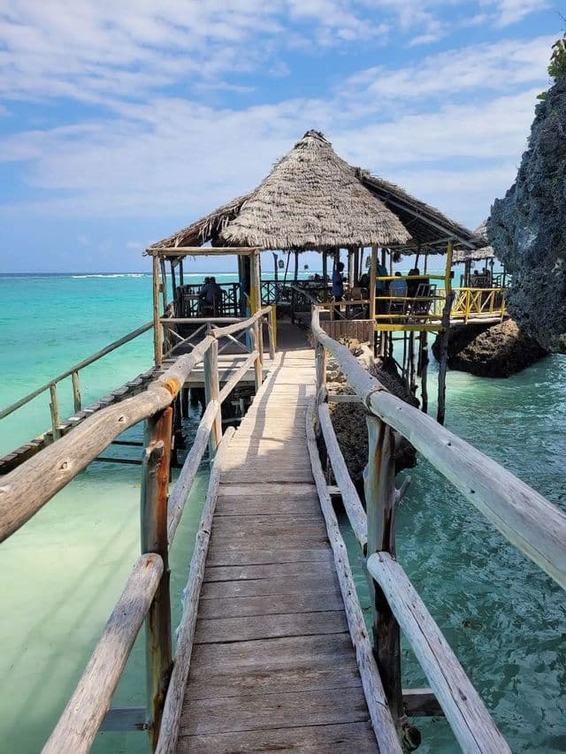 Une passerelle en bois avec des balustrades rustiques menant à un restaurant au toit de chaume construit sur pilotis au-dessus d'une eau turquoise.